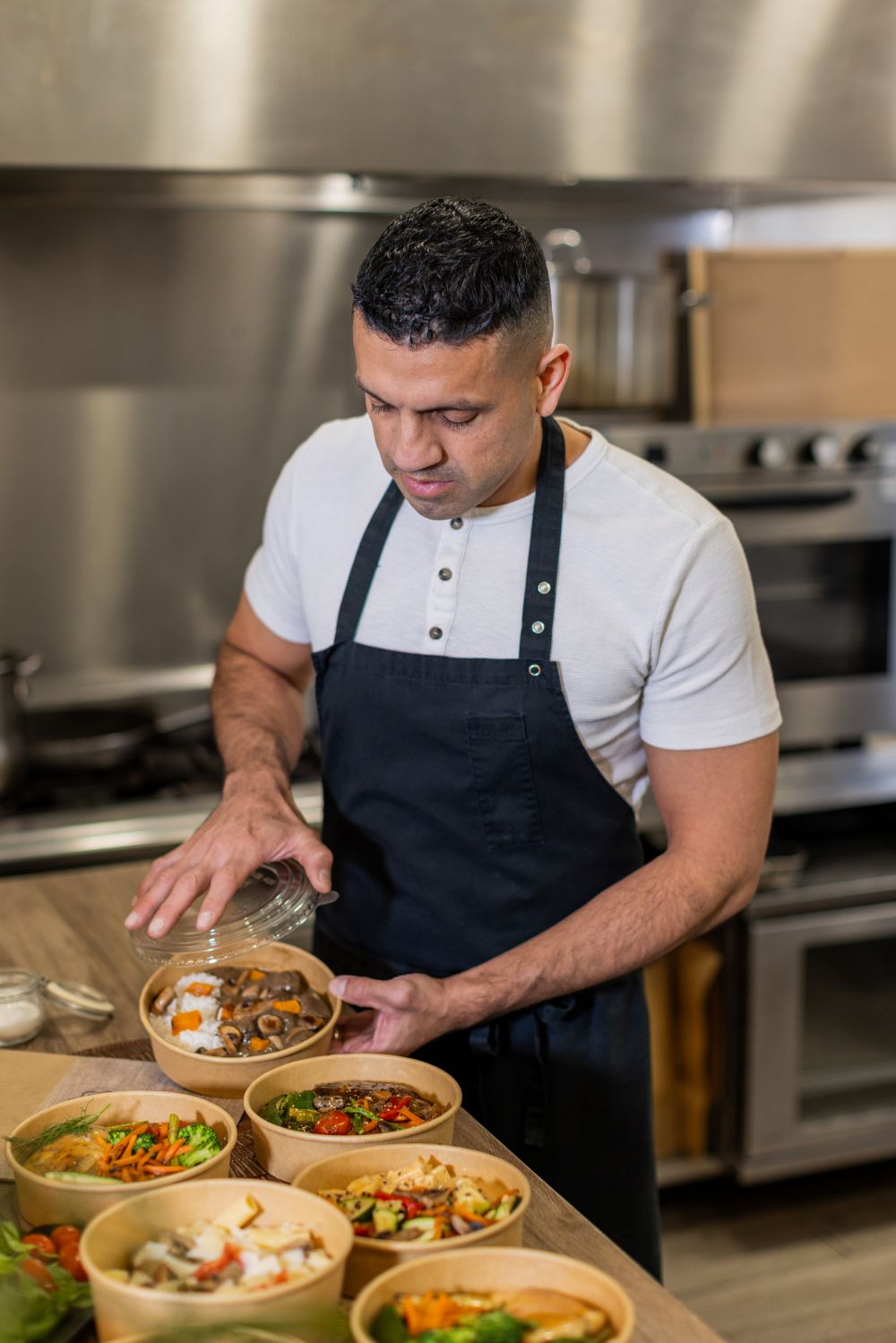 A chef in a black apron packs fresh, healthy takeout meals in eco-friendly bowls. A behind-the-scenes look at a professional kitchen focused on sustainable food service