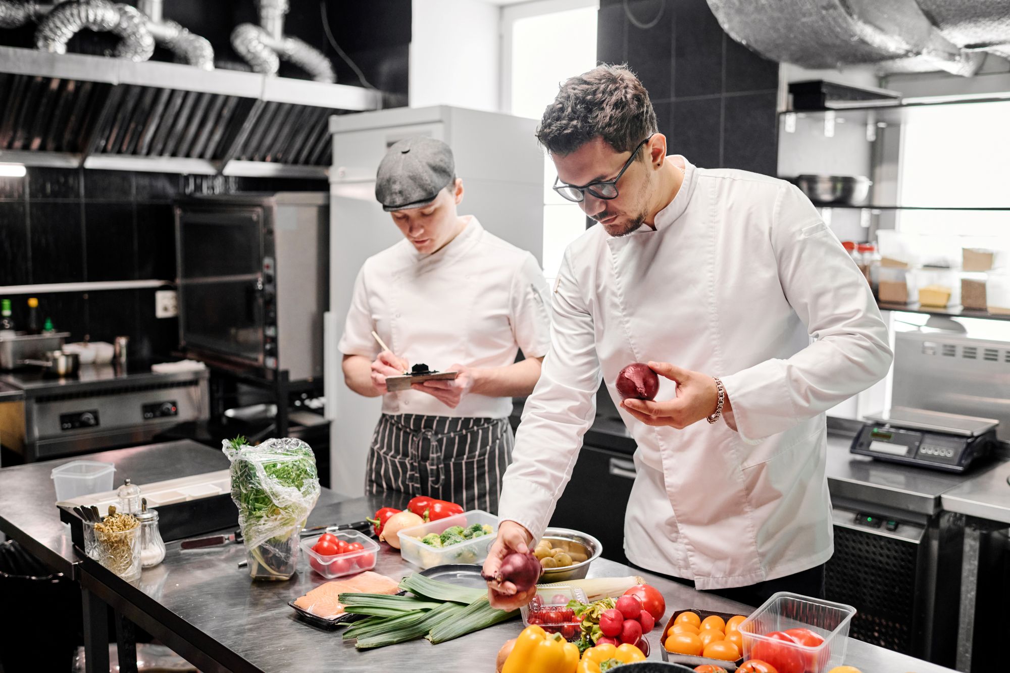 Young chef in uniform choosing fresh vegetables for dish at table with his assistant making notes on paper, they working in commercial kitchen